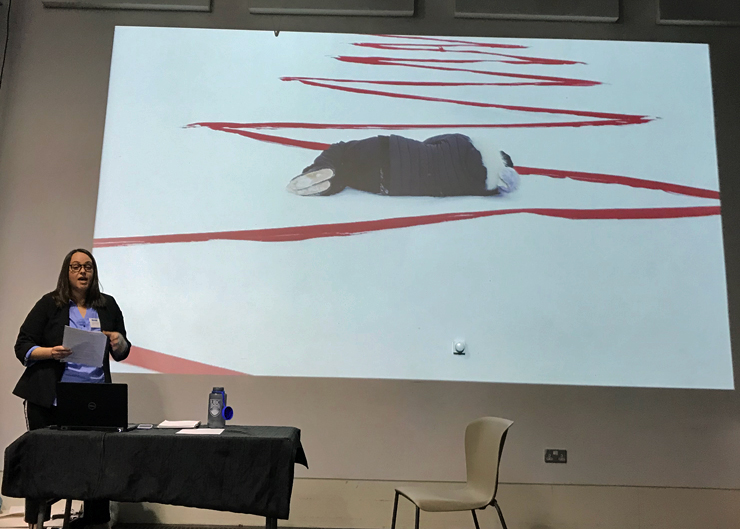Woman speaking in front of a slide depicting a person lying on the snow-covered ground within a red zigzag line.